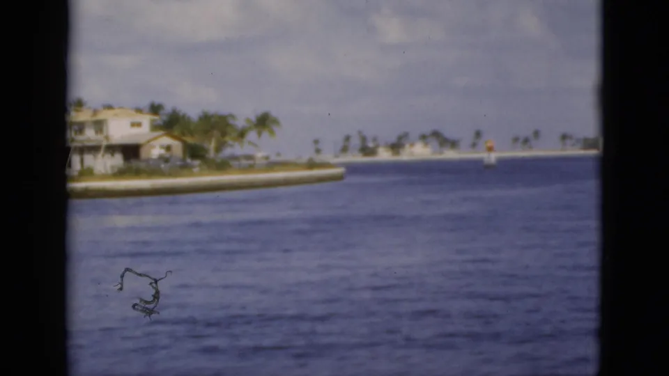 1975 Florida 8mm frame looking across open water toward low shoreline buildings and palms under a partly cloudy sky.