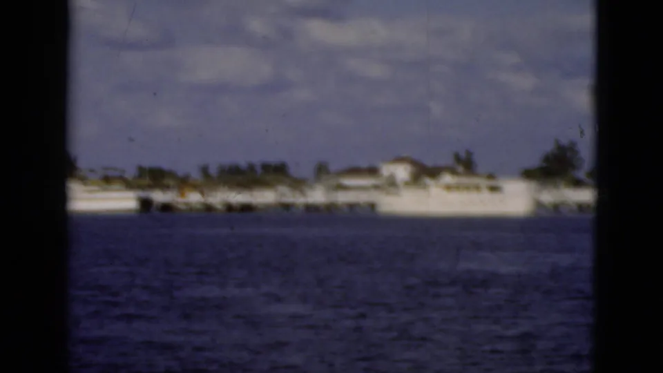 Soft 1975 Florida 8mm view of distant waterfront buildings across the water, with cloudy sky and dark frame borders.