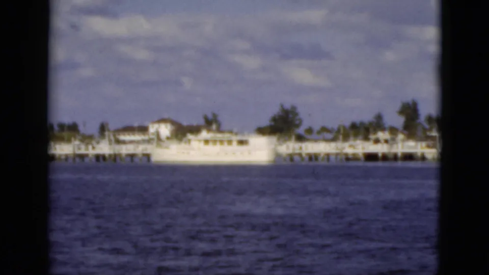 Soft 8mm view of a Florida waterfront dock and white vessel across blue water, with shoreline trees and clouds in the background.