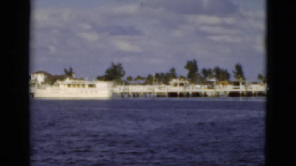 1975 Florida home-movie frame with a long white dock, shoreline structures, palms, and blue water under a bright cloudy sky.