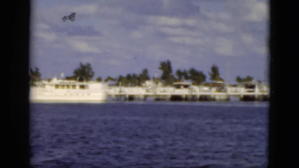 Florida 1975 8mm frame showing dock pilings, low waterfront structures, trees, and rippled blue water from a distant viewpoint.