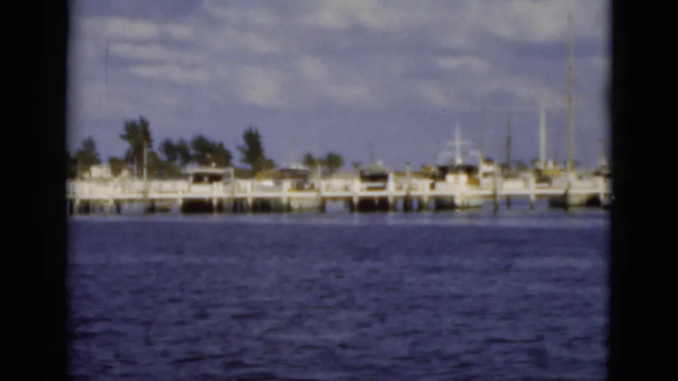 1975 Florida waterfront scene with docked boats, pier structures, palms, and calm blue water viewed from offshore.