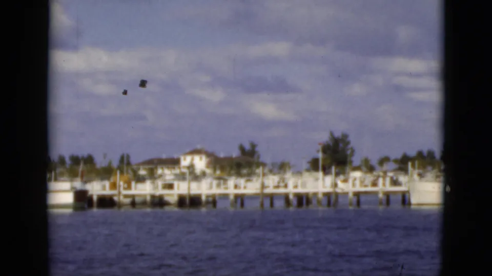 1975 Florida waterfront view with a pier, small buildings, trees, and boats across blue water, with visible 8mm film marks.