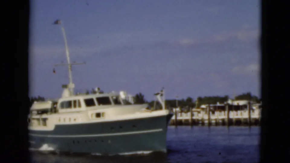 1975 Florida 8mm frame showing a white cabin motor yacht crossing calm water beside dock structures.