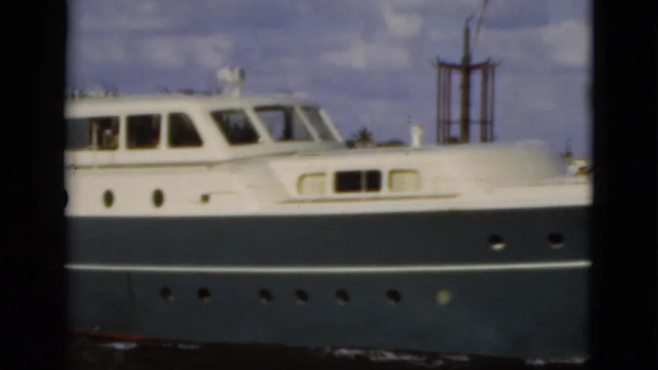 Close 1975 Florida home-movie view of a white and blue motor yacht side passing near a waterfront dock.