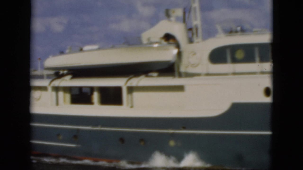 1975 Florida 8mm frame of the yacht midsection, showing a light-colored dinghy strapped above the cabin and portholes along the blue hull.
