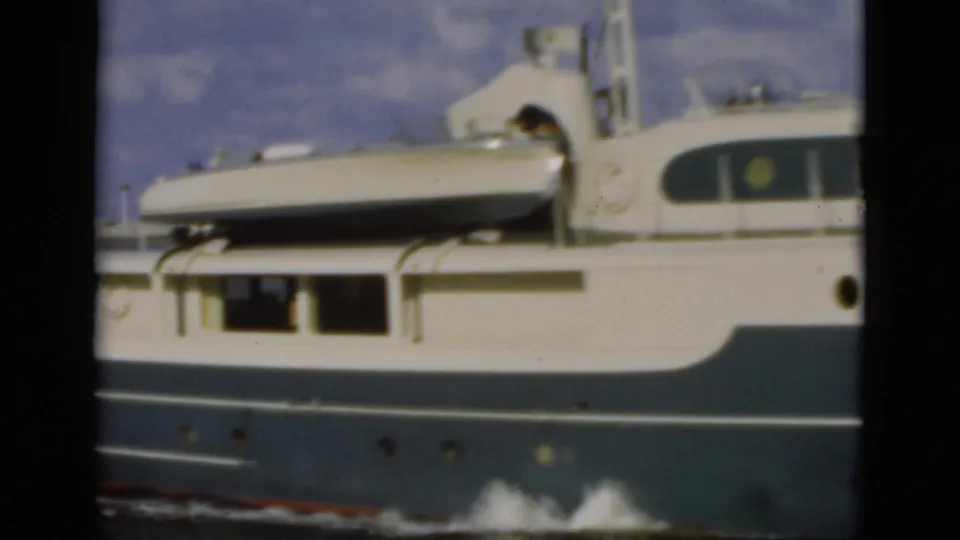 1975 Florida 8mm frame of the yacht midsection, showing a light-colored dinghy strapped above the cabin and portholes along the blue hull.