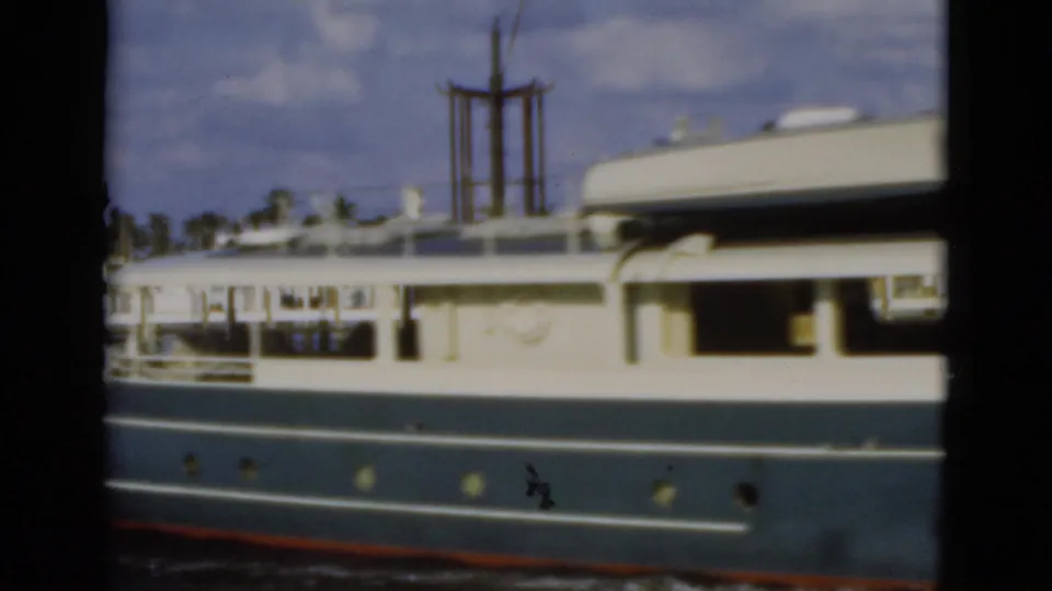 1975 Florida 8mm frame showing the yacht broadside with a dinghy on deck, railings, portholes, and faint shoreline vegetation beyond the vessel.