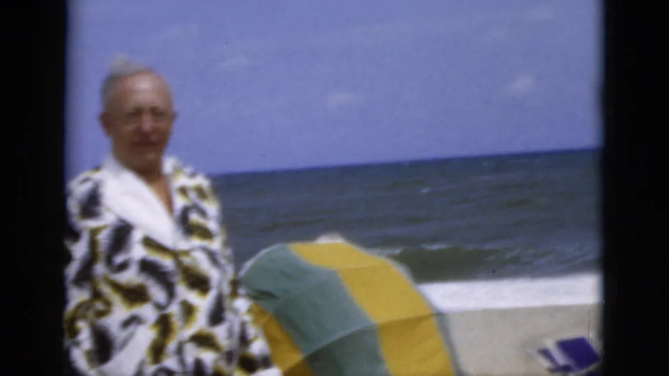 Older man on a Florida beach in 1975 stands beside a green and yellow striped umbrella, with surf and ocean horizon behind him.