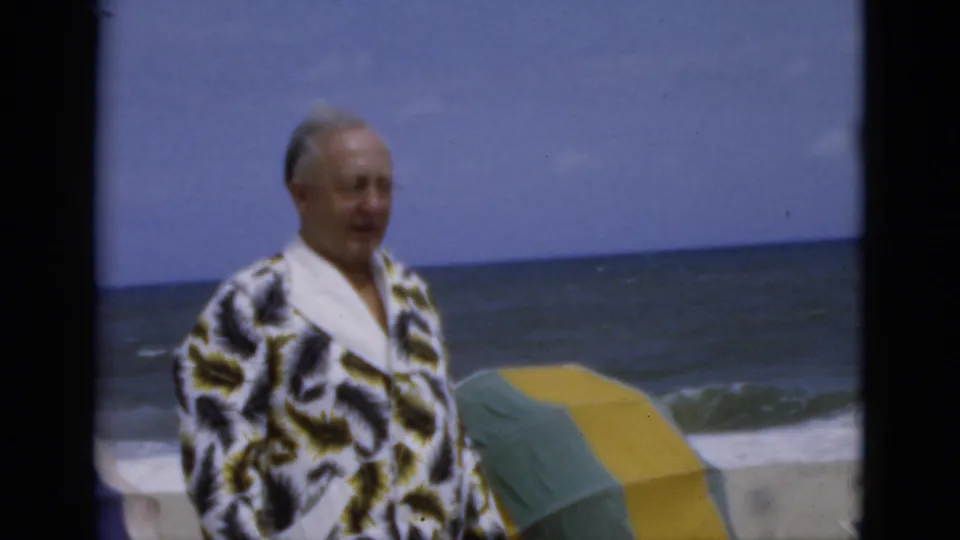 1975 Florida beach frame of an older man in a patterned robe beside a green and yellow umbrella, with waves and horizon visible.
