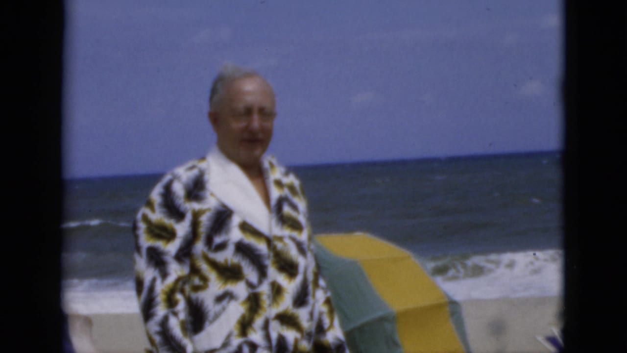 Adult man in a patterned dressing gown stands on a beach with ocean waves and a striped umbrella behind him.