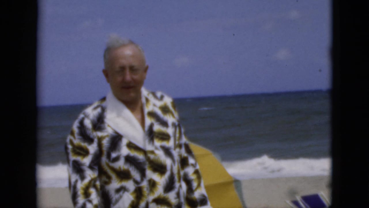 Adult man in a patterned dressing gown faces the camera beside a striped beach umbrella and rough ocean.