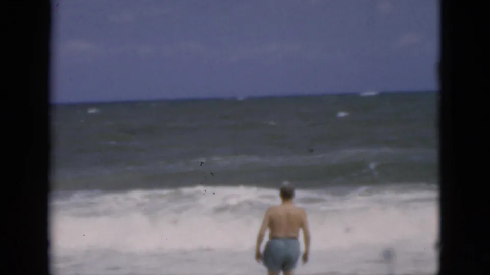 In the same 1975 Florida beach scene, the man is shirtless in blue trunks and walks toward rough, foaming waves under a cloudy sky.