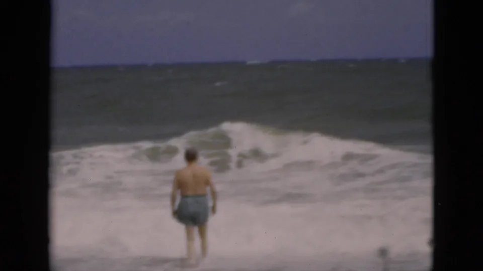 In 1975 Florida beach footage, a middle-aged man in blue trunks faces churning surf under a hazy sky.