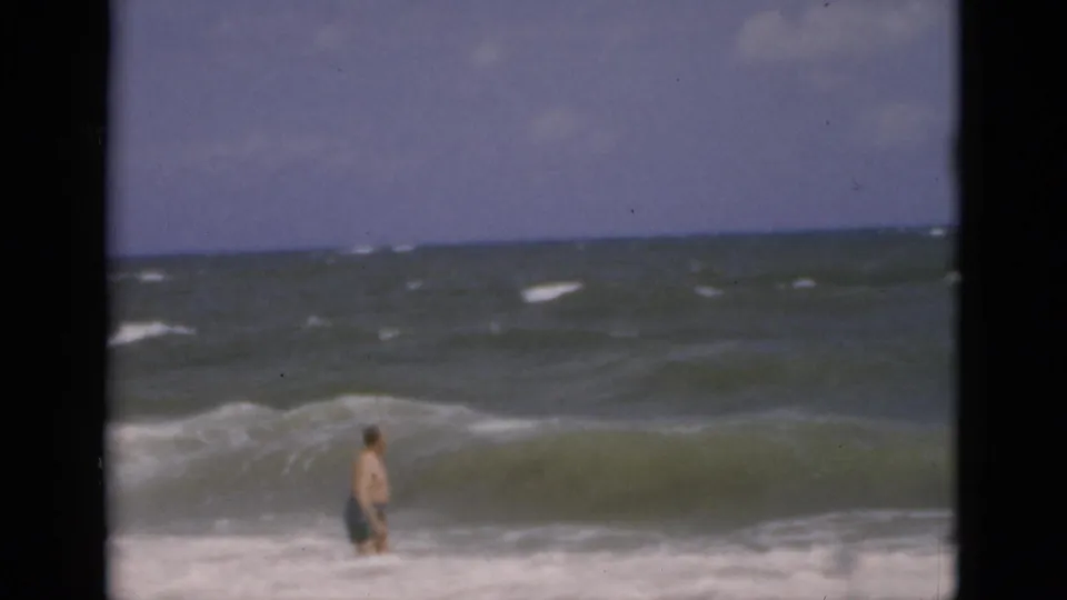 A distant adult man wades into Florida surf as a green wave begins to crest in the 8mm frame.