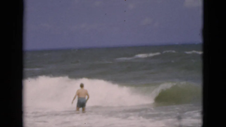 A middle-aged man in blue trunks faces the ocean as white surf spreads across the Florida beach scene.