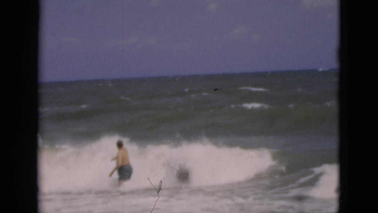 Male bather in blue trunks walking into foamy surf under a hazy sky.
