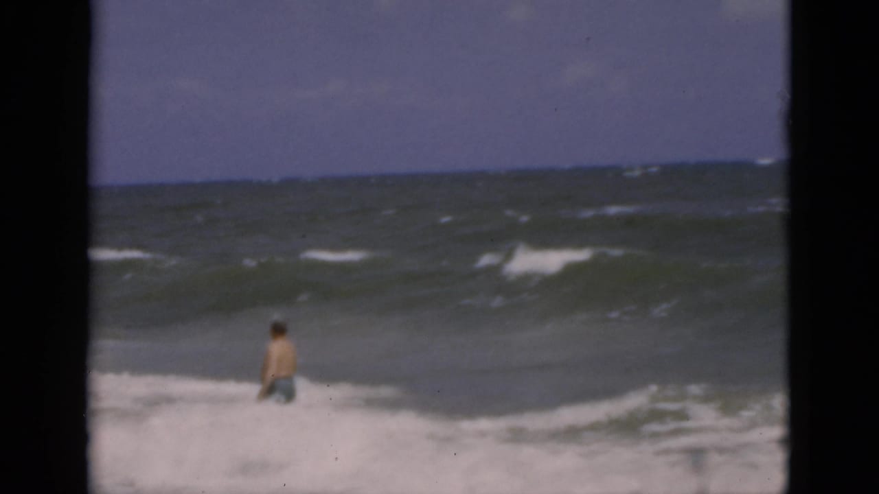 1975 Florida beach frame with a male bather waist-deep in surf, seen from shore across choppy water and a hazy horizon.