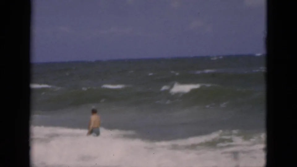 1975 Florida beach frame with a male bather waist-deep in surf, seen from shore across choppy water and a hazy horizon.