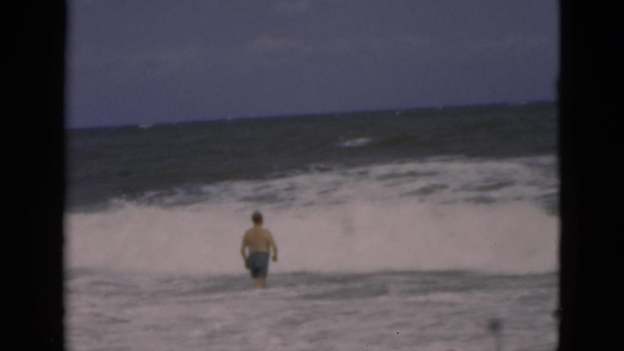 Shirtless man in swim trunks stands in ocean surf as a wave breaks behind him.