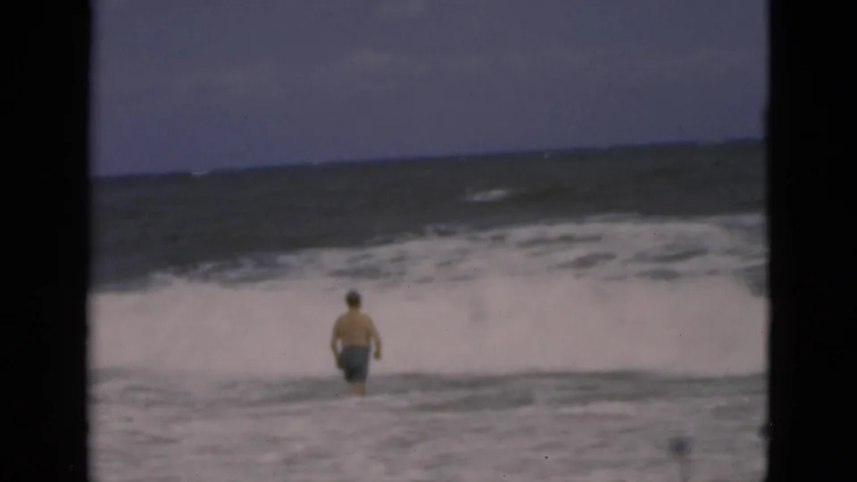 In supported 1975 Florida beach footage, a shirtless man stands in churning surf with white waves and a dark ocean horizon behind him.
