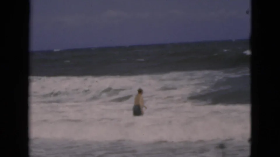 A shirtless man in swim trunks stands low in the frame as white surf crosses the Florida beach scene and the ocean horizon remains visible.