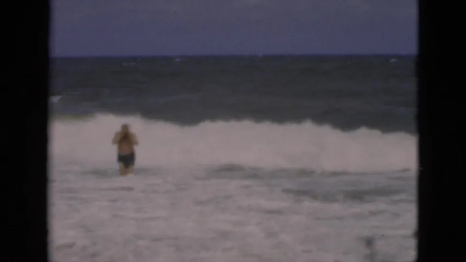 1975 Florida beach home-movie frame showing a man standing in shallow surf as white waves break offshore.