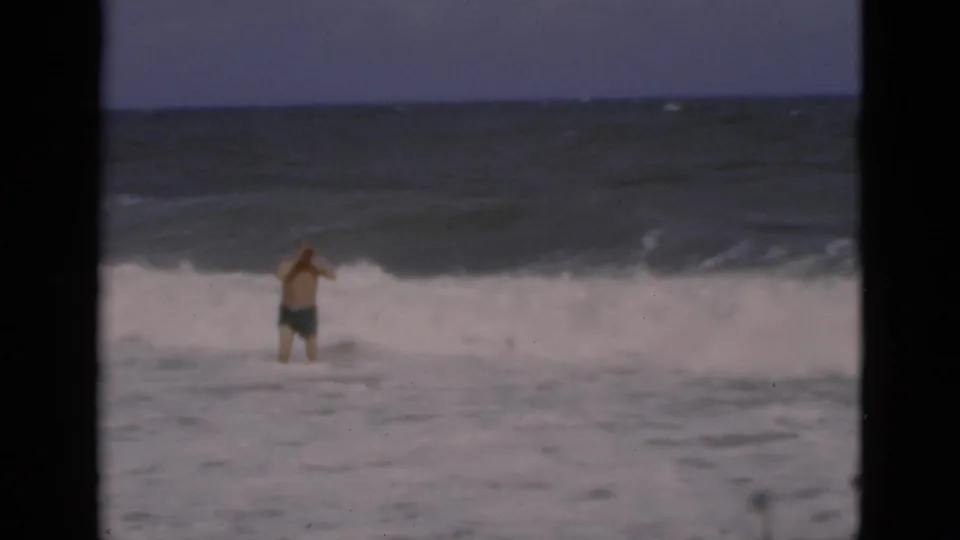 A man in swim trunks wades through foamy Florida surf in soft, grainy 8mm home-movie footage.