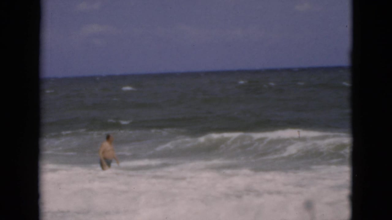 Man standing waist-deep in rough surf with whitecaps and a cloudy horizon behind him.