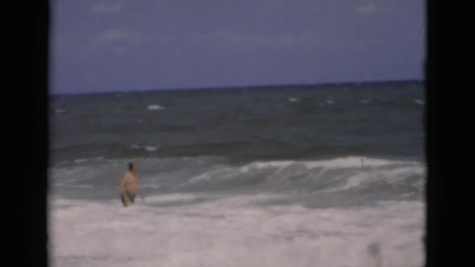 A man in swim trunks stands in the churning ocean at a Florida beach, framed by dark vertical bars in 1975 8mm footage.
