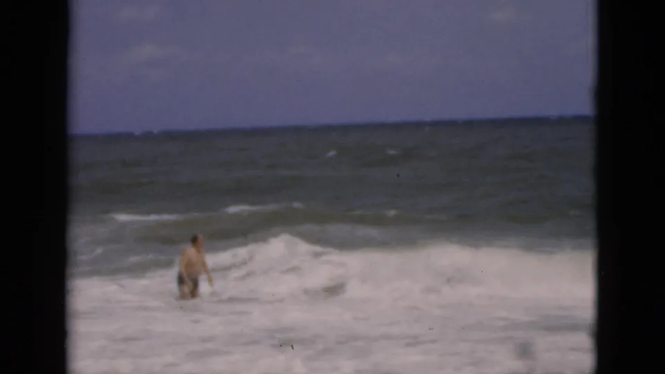 The swimmer wades through foamy Florida surf while a breaking wave rises beside him in soft 1975 8mm color footage.