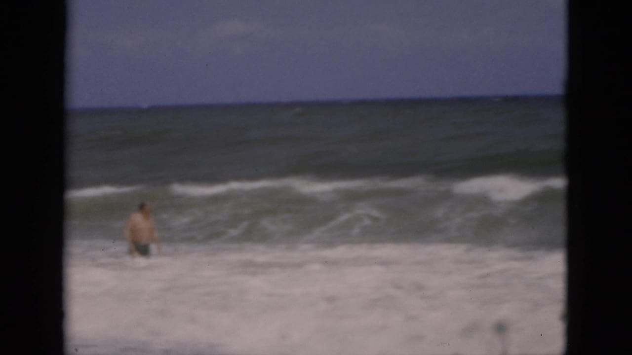 1975 Florida 8mm frame showing an adult man wading in foamy ocean surf beneath a muted sky.