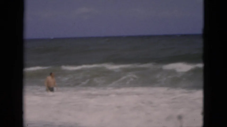 1975 Florida 8mm frame showing an adult man wading in foamy ocean surf beneath a muted sky.