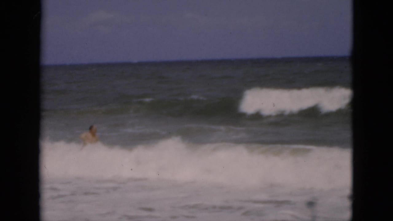 1975 Florida beach frame with a distant adult swimmer among white breaking waves and dark ocean water.
