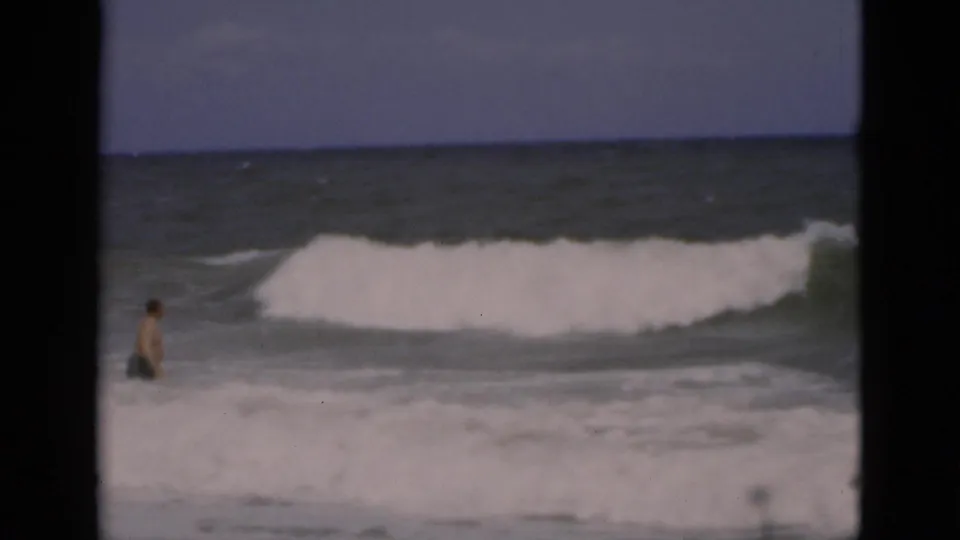 1975 Florida 8mm frame showing a man wading at the left edge of foamy surf as a larger wave rolls behind him.