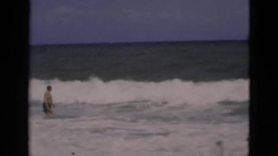 1975 Florida beach frame with an adult man walking through shallow surf as a white wave breaks across the background.