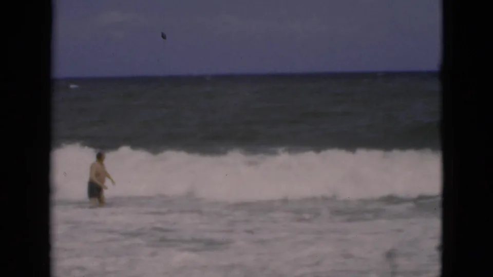 In 1975 Florida home-movie footage, a man in swim trunks wades through foamy ocean surf under a blue sky.