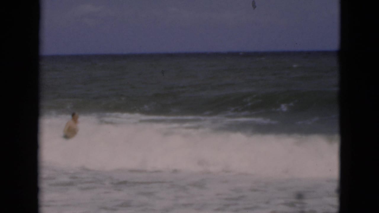 A man wades into churning Florida surf in grainy 1975 8mm footage, seen between dark vertical frame bars.