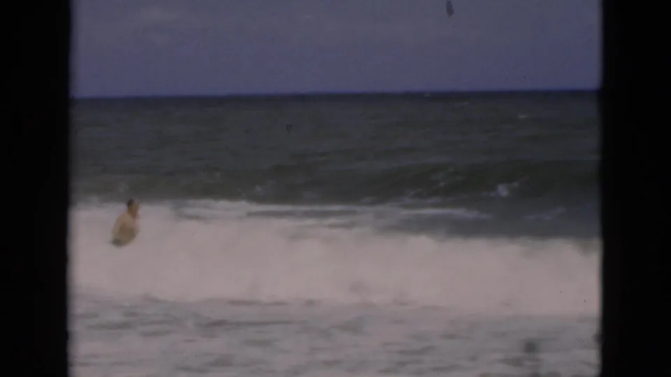 A man wades into churning Florida surf in grainy 1975 8mm footage, seen between dark vertical frame bars.