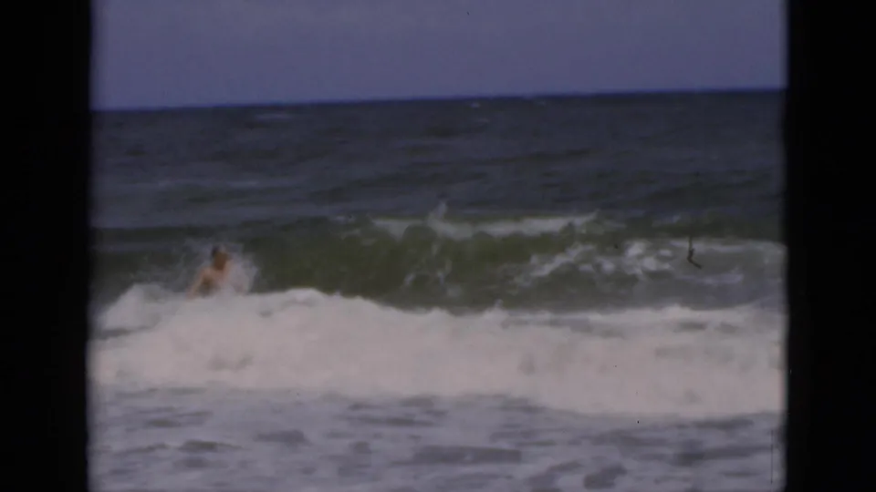 The man is low in foamy surf while a dark green wave crests behind him in the 1975 Florida beach footage.