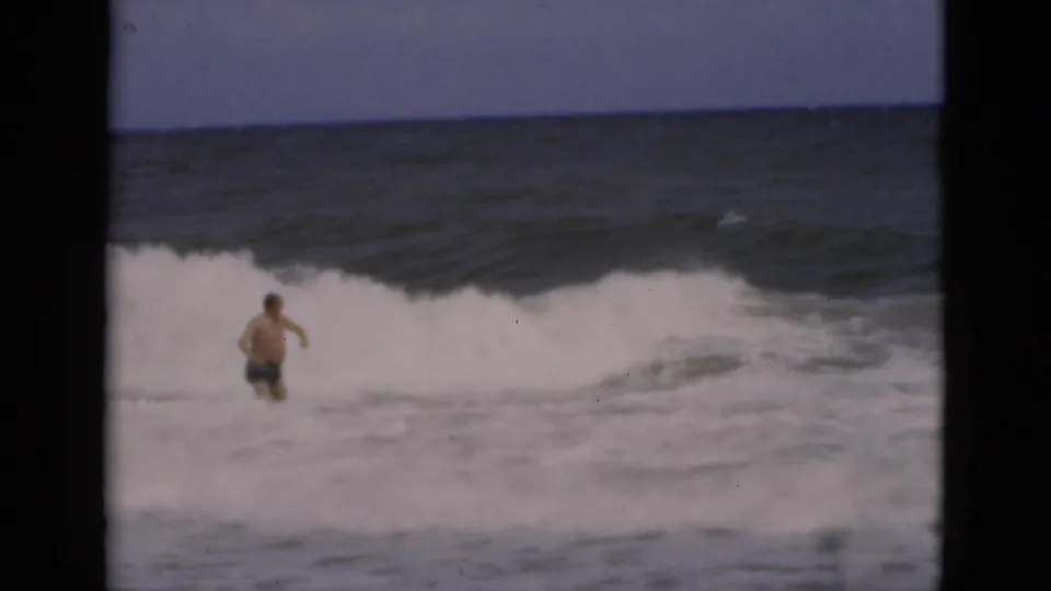 A man seen from behind stands in foamy Florida surf as a wave breaks ahead of him in soft 1975 8mm footage.
