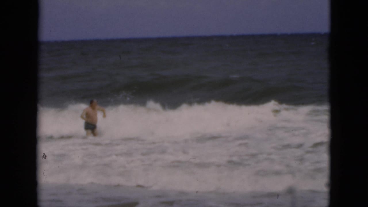 Shirtless man wading in rough ocean surf near breaking waves.