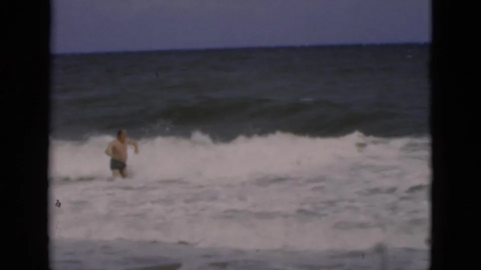 A shirtless adult man in dark swim trunks wades through heavy surf at a Florida beach in 1975 home-movie footage.
