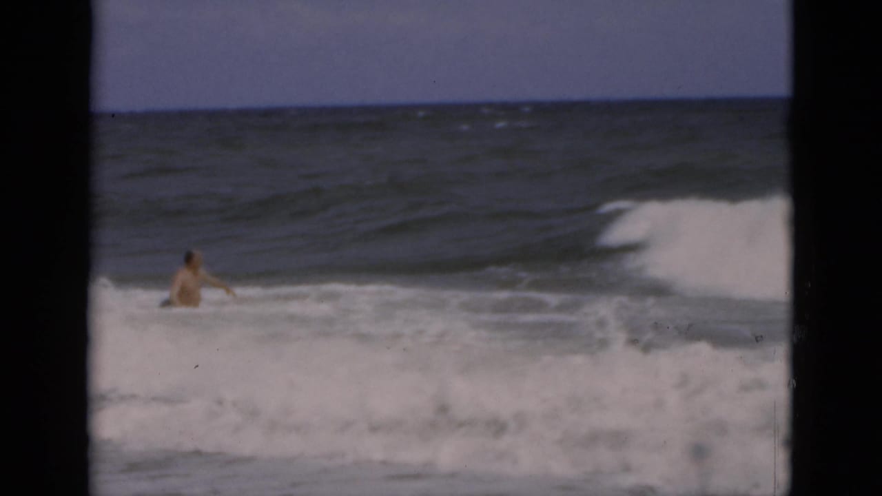 Adult man wades in foamy ocean surf with a breaking wave behind him.