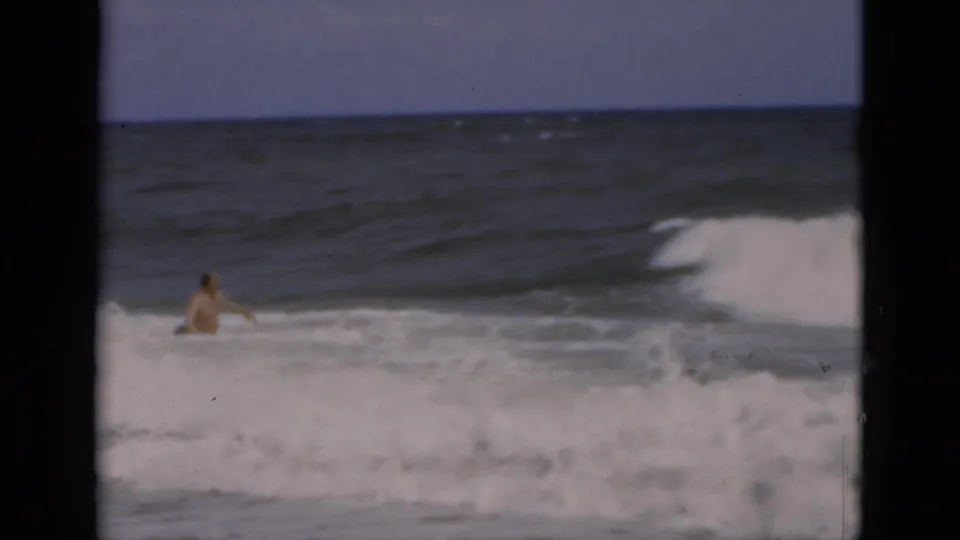 1975 Florida 8mm frame of an adult man standing in white surf as a wave breaks across the ocean behind him.