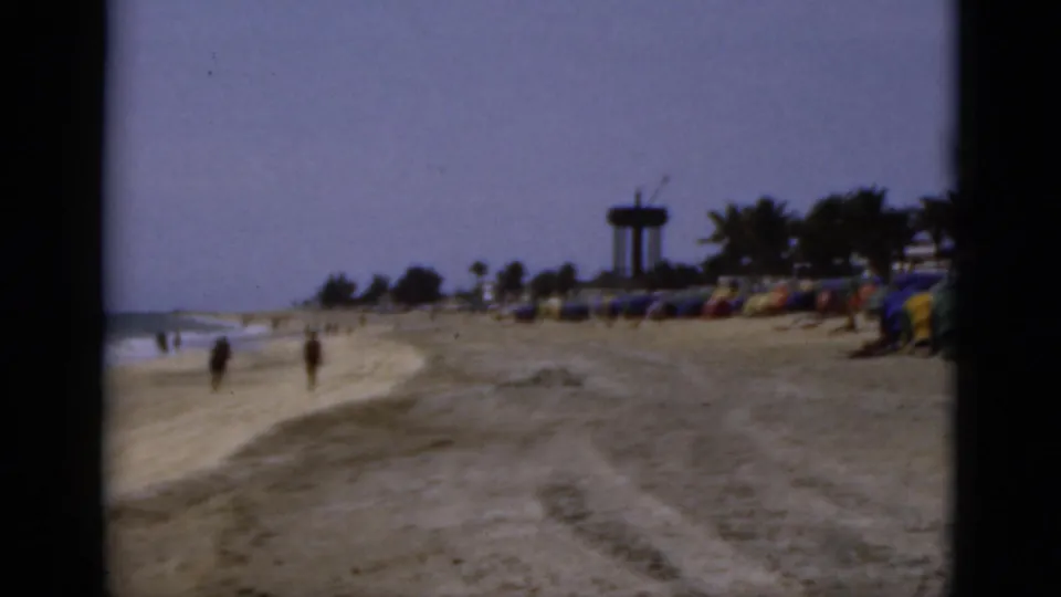 1975 Florida 8mm view down a sandy beach with surf at left, pedestrians, umbrella rows, and a distant water-tower-like structure.