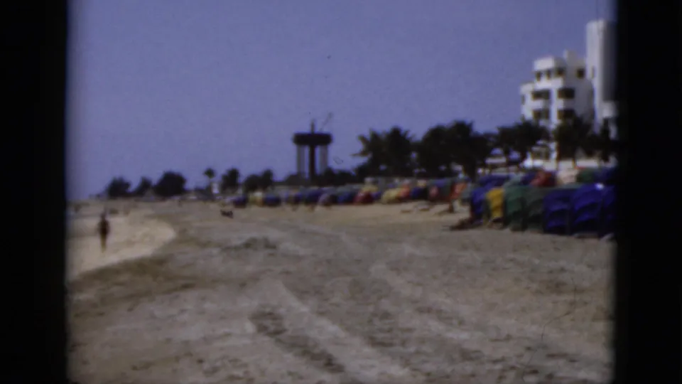 1975 Florida beach frame with rows of umbrellas, scattered beachgoers, distant tower, palm trees, and beachfront buildings.
