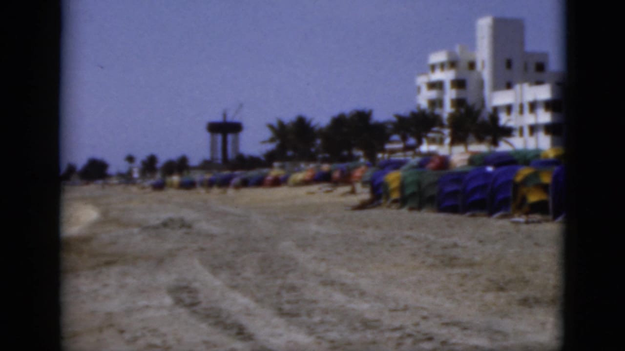 1975 Florida beach view with rows of colorful shade tents, white seaside buildings, palm trees, and a distant tower-like structure.