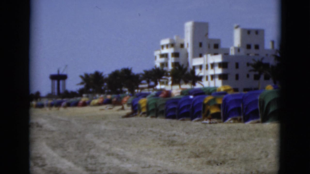 1975 Florida shoreline scene with low beach tents, white Art Deco-style coastal buildings, palms, parked cars, and open sky.