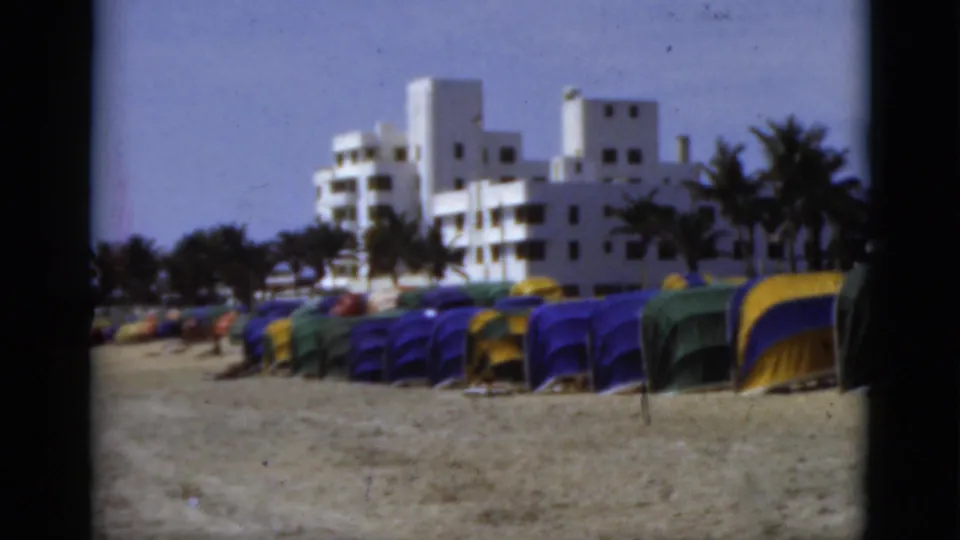 1975 Florida beach frame showing colorful tents in the foreground, white coastal buildings, palms, and a distant figure near the tents.